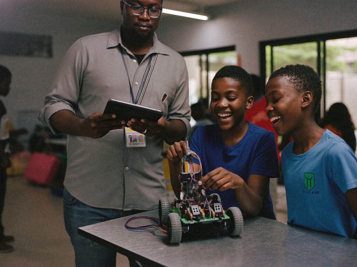 Two learners smile while wiring a small robotics kit on a workbench as a supervising facilitator holds a tablet.