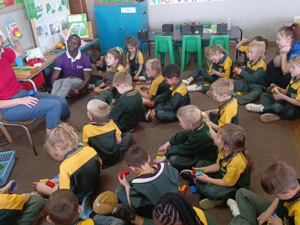 A teacher holds up a brick stack while about twenty-five uniformed pupils sit on the floor, each with coloured bricks in hand