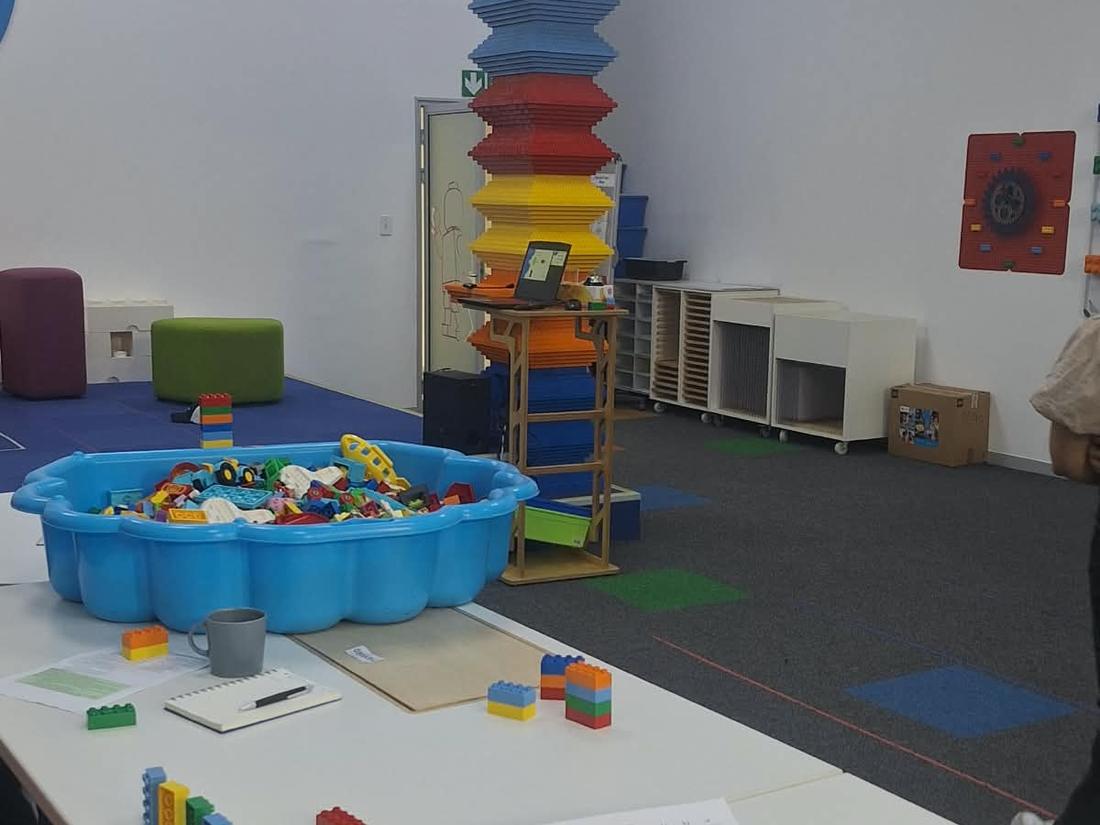 A play and training room with a screen reading Can we code, showing brick icons for move forward, turn and stop, and tables set up with brick programs