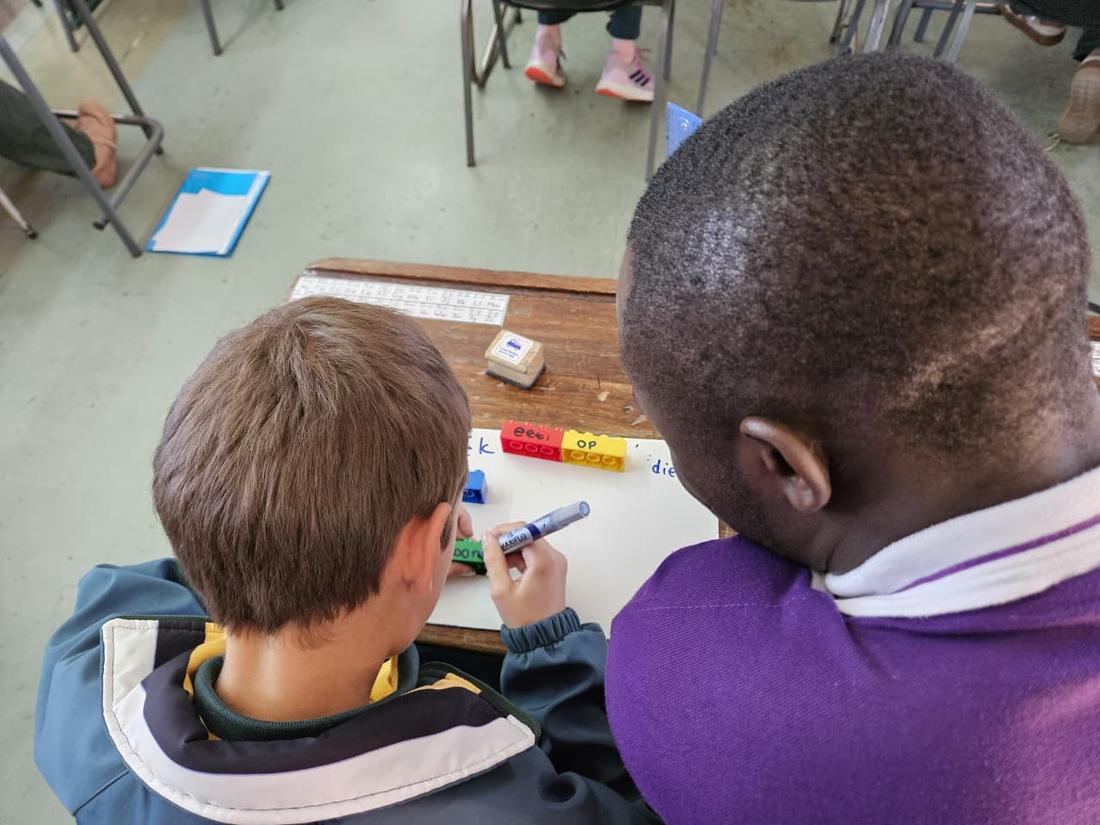 A facilitator leans over a pupil writing on a mini whiteboard, word bricks on the desk between them