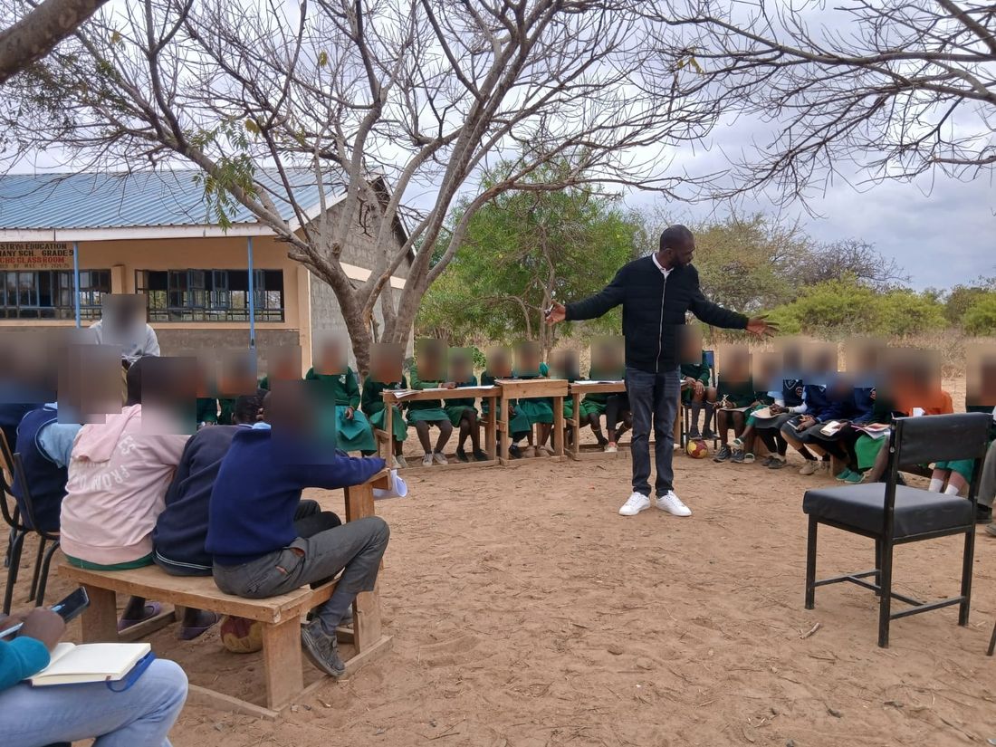 A facilitator with arms open leads a large circle of uniformed pupils seated on benches under an acacia tree outside a rural Kenyan school; pupils' faces are blurred