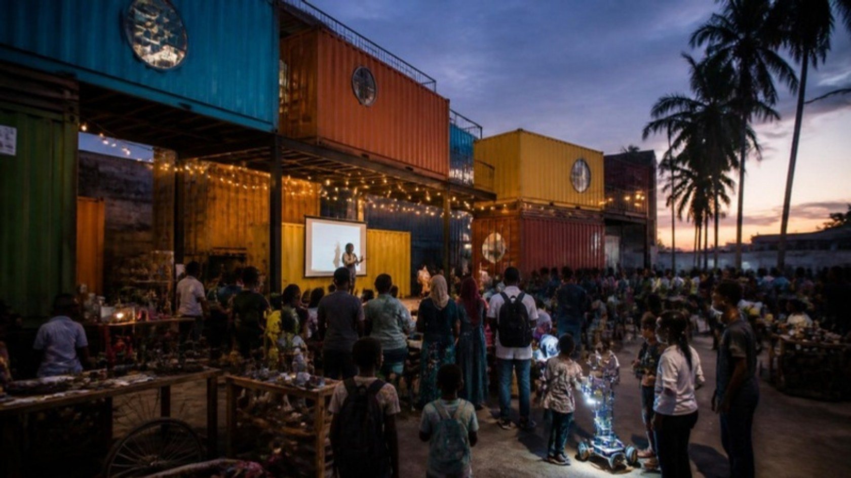 Concept render: an evening fair under string lights beneath the stacked containers, with stalls and a small stage
